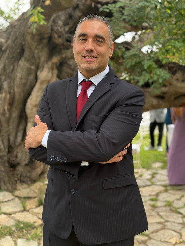 Mann im dunklen Anzug mit roter Krawatte vor einem großen Baum im Außenbereich