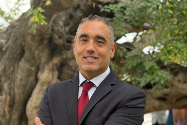 Mann im dunklen Anzug mit roter Krawatte vor einem großen Baum im Außenbereich