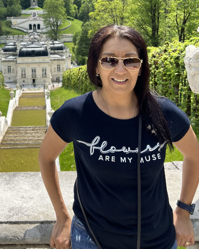 Frau mit Sonnenbrille in dunkelblauem T-Shirt vor einer historischen Parkanlage mit Treppen und Gebäuden