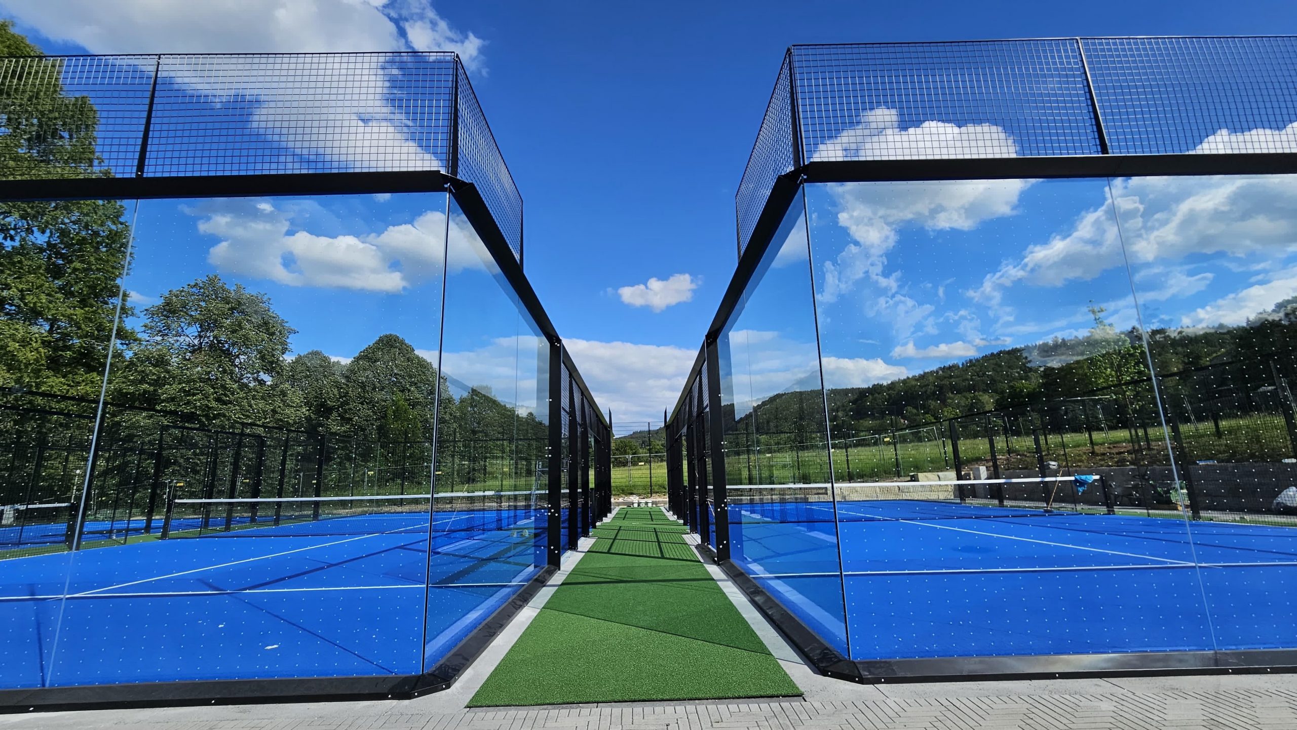 Zentraler Verbindungsweg zwischen zwei Outdoor-Padelplätzen mit Glaswänden und blauem Spielbelag