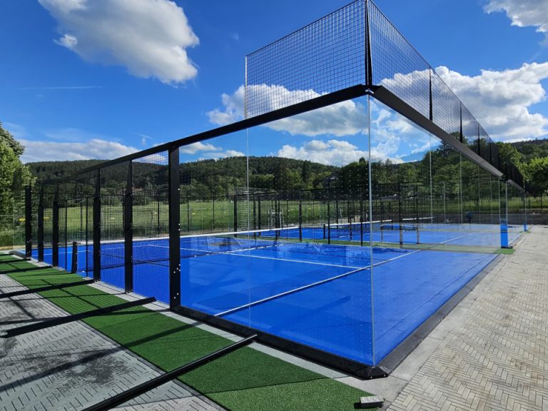 Outdoor-Padelplatz mit Glaswänden, schwarzer Stahlkonstruktion und blauem Spielbelag unter blauem Himmel