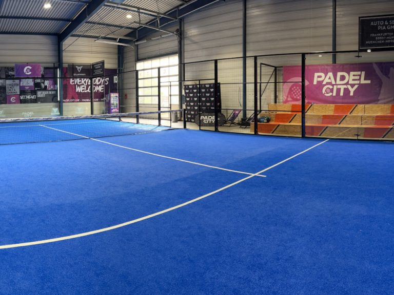 Indoor-Padelplatz mit blauem Spielbelag, Glaswänden und Zuschauerbereich