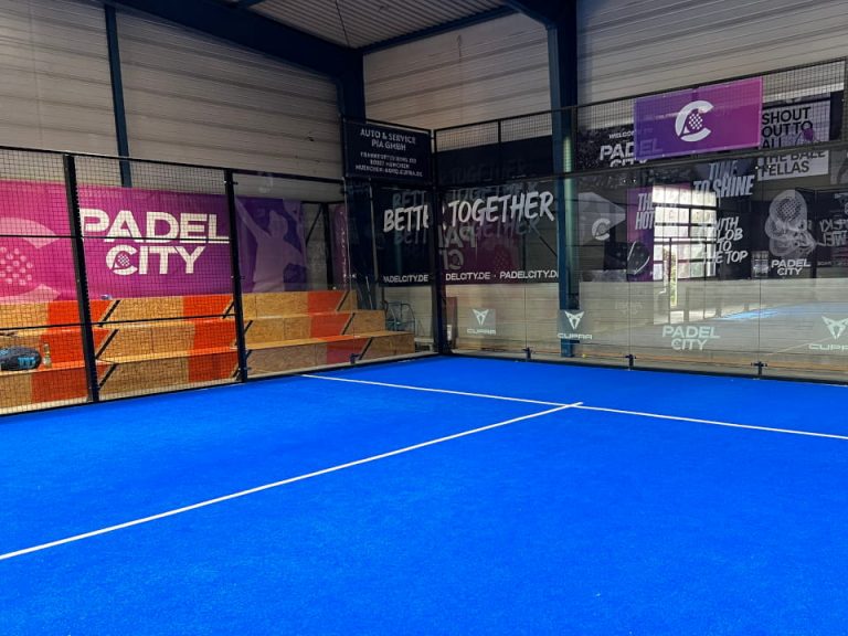 Indoor-Padelplatz mit blauem Spielbelag, Glaswänden und Tribünenbereicht