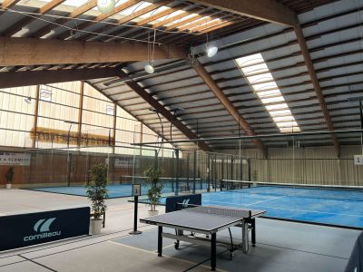 Seitliche Ansicht einer Indoor-Padelhalle mit blauen Padelplätzen, Glaswänden und einem Tischtennistisch im Vordergrund.